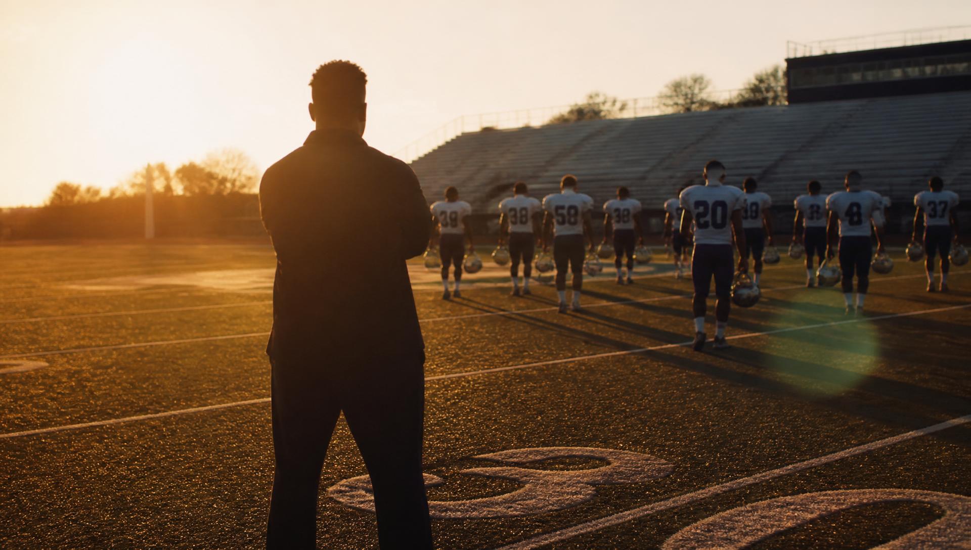 A coach standing at midfield watching athletes walk off the field at sunset