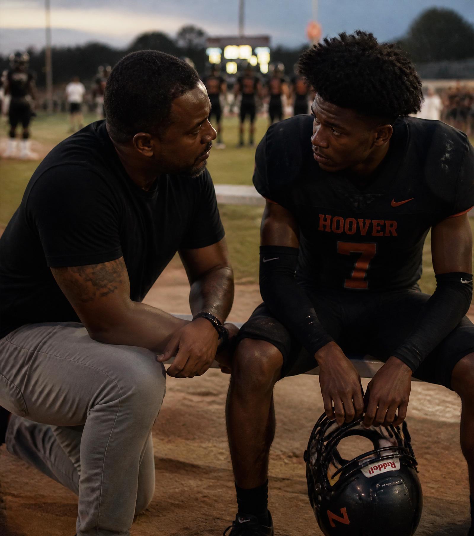A father kneeling beside his teenage son at the edge of a soccer field at dusk — quiet presence, no fixing