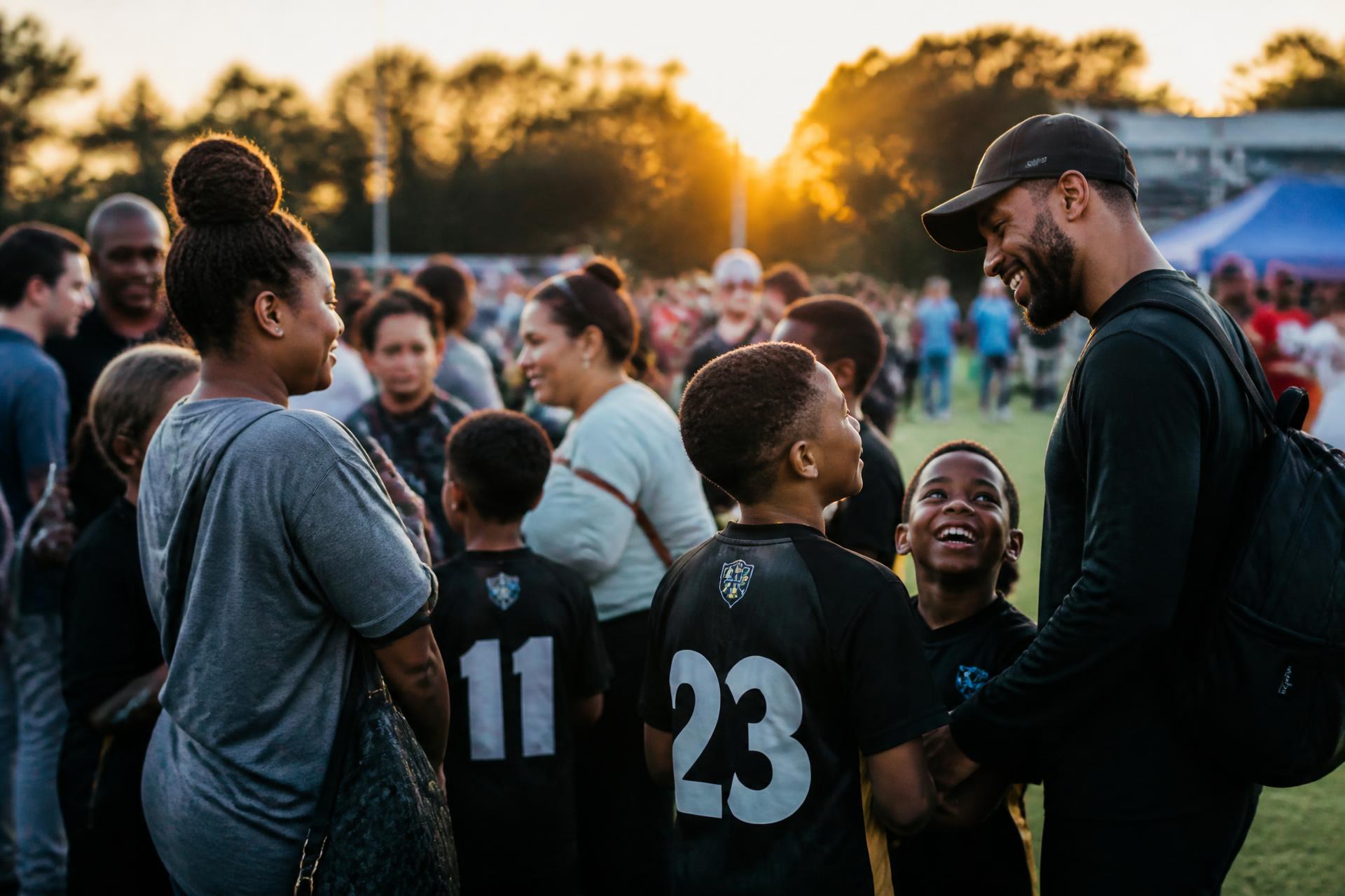 A diverse community of families gathering after a youth sports event at golden hour — belonging, community, hope