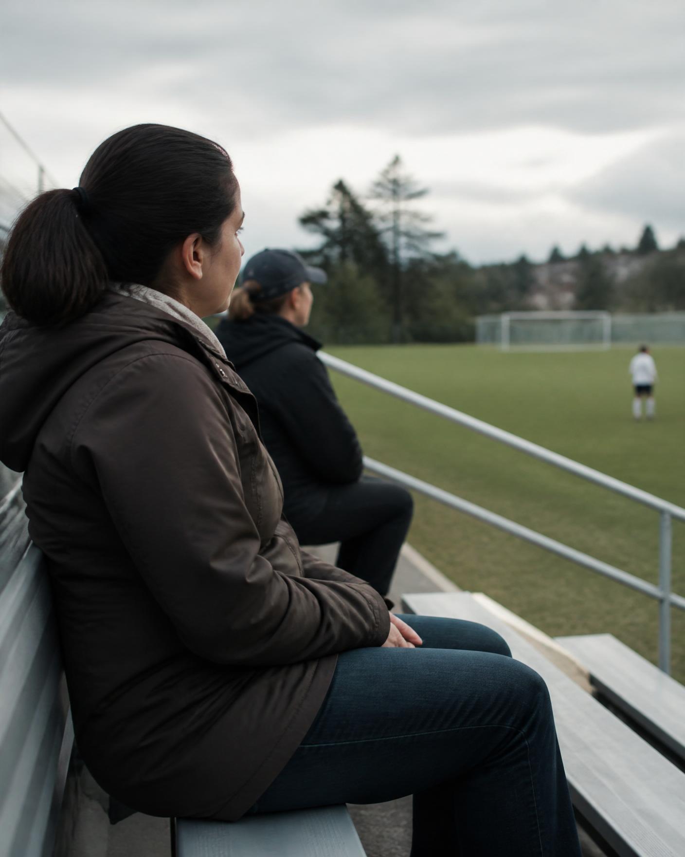 A parent sitting quietly in the stands, hands folded, watching a youth soccer game from the distance