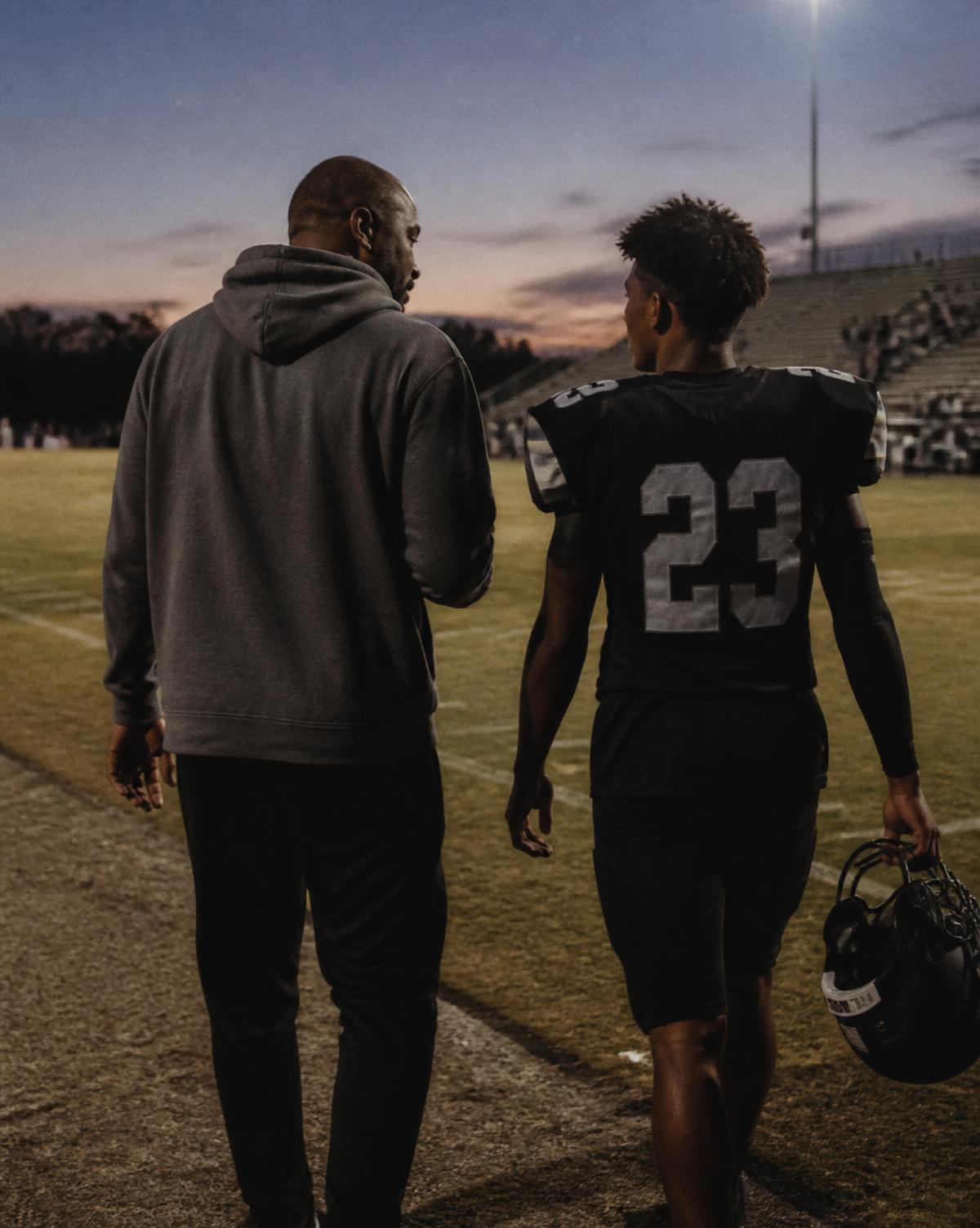 A Black father and his teenage son in a football jersey walking off the field together at dusk