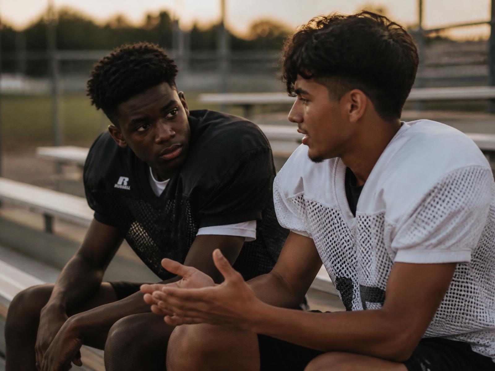 Two teammates in practice jerseys sitting on empty bleachers having a genuine conversation after practice