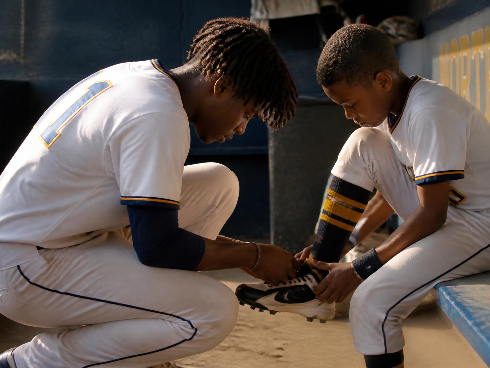 Older baseball athlete crouched down helping a younger teammate in the dugout — quiet mentorship