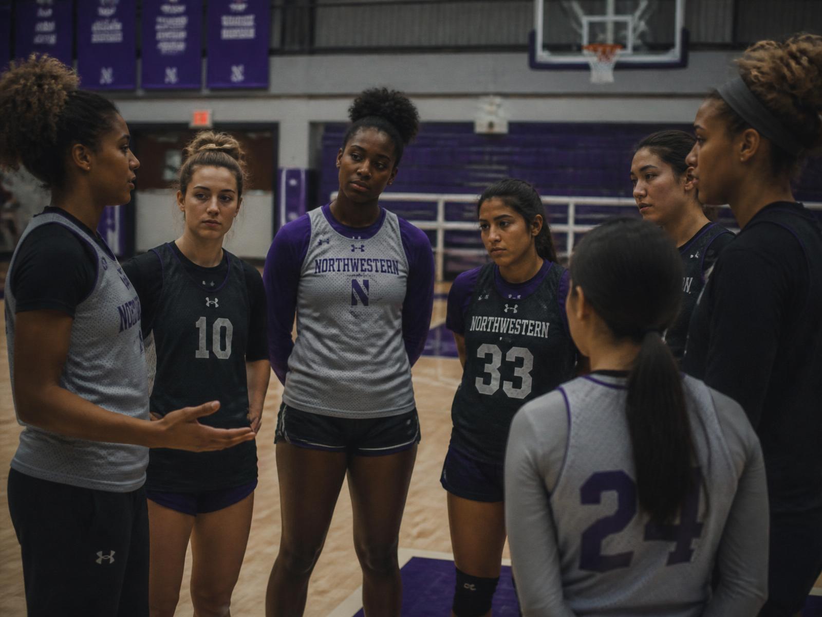 Female basketball team standing in a quiet circle on the court after practice, listening to one teammate