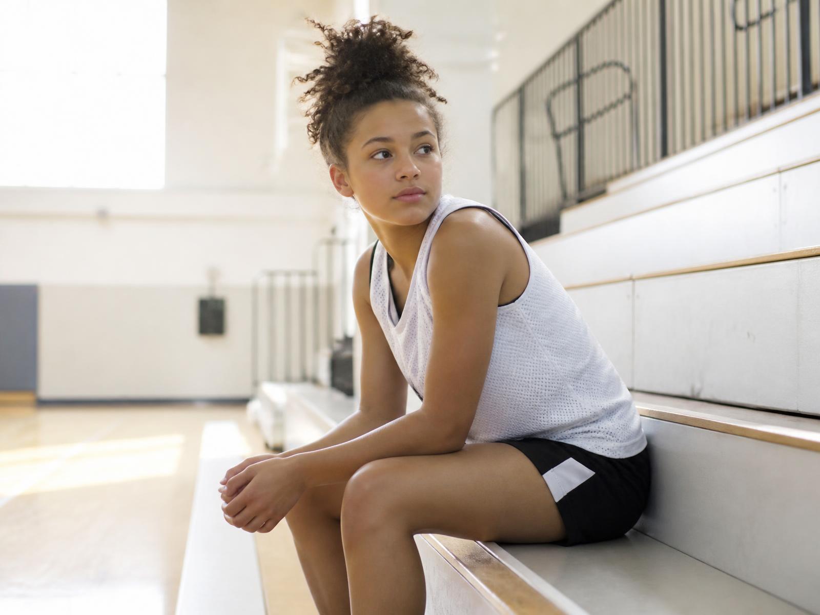 A teenage girl athlete sitting alone on bleachers in a sunlit gym between moments